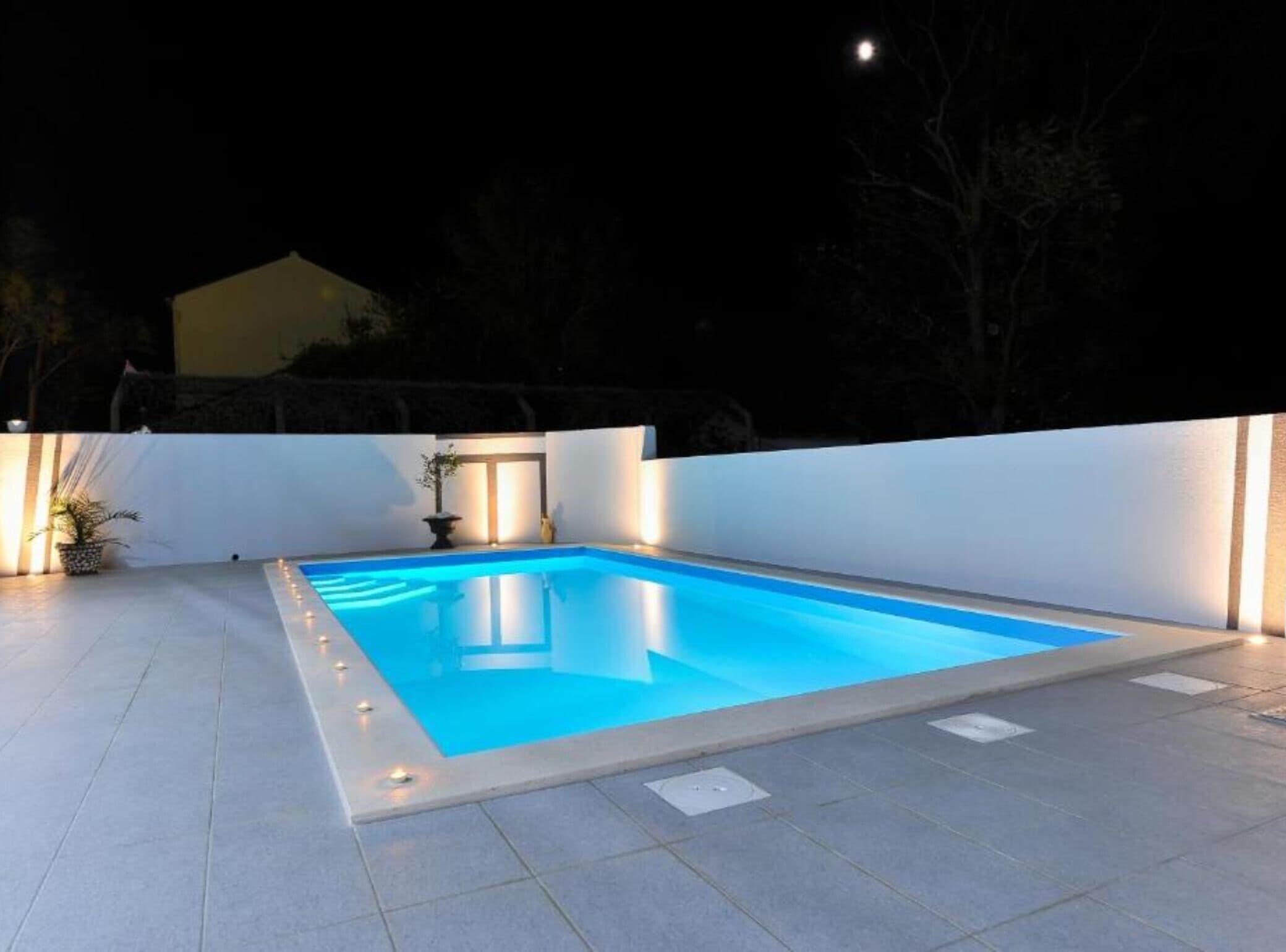 A rectangular swimming pool with a light blue interior, surrounded by a white wall and a tiled deck.