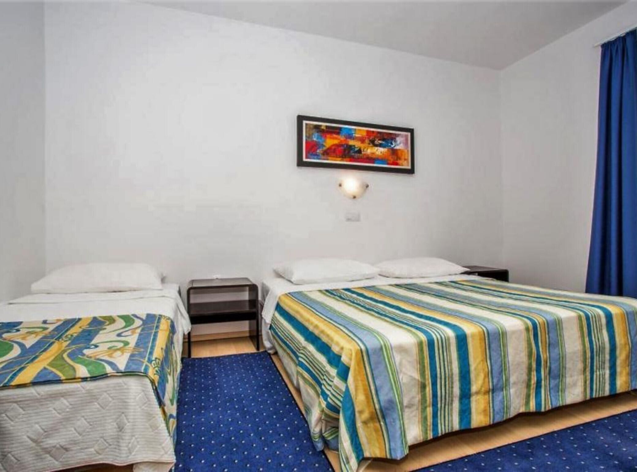 Camera d'albergo con due letti, un tavolino, una lampada a parete e un quadro astratto dai colori vivaci.