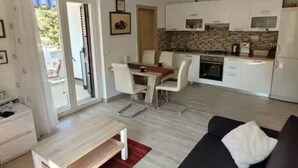 Eine moderne Küche mit weißen Schränken, einer Mikrowelle und einem Herd. Ein Essbereich mit Tisch und Stühlen ist vorhanden. Im Wohnbereich befinden sich eine schwarze Couch und ein kleiner Tisch mit einem roten Teppich.
