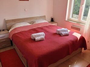 Una cama bien hecha con una colcha roja y dos toallas blancas dobladas, una mesita de noche con una lámpara y una ventana con vistas a zonas verdes.