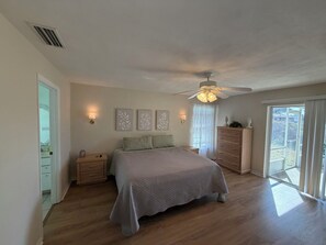 Une chambre à coucher comprenant un lit, des tables de chevet, une commode, un ventilateur de plafond et une vue sur l’extérieur, visible à travers une porte vitrée.