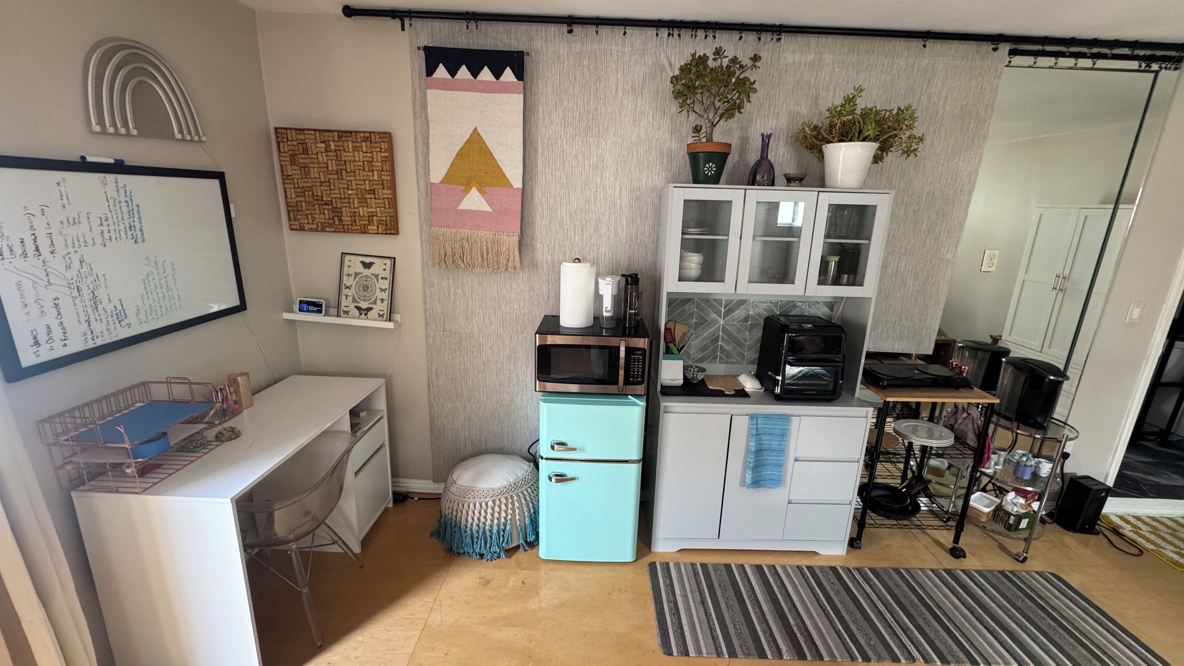 Cocina compacta con microondas, tostadora y refrigerador pequeño. Hay una pizarra con notas, un escritorio pequeño con silla y un tapete a rayas en el suelo.