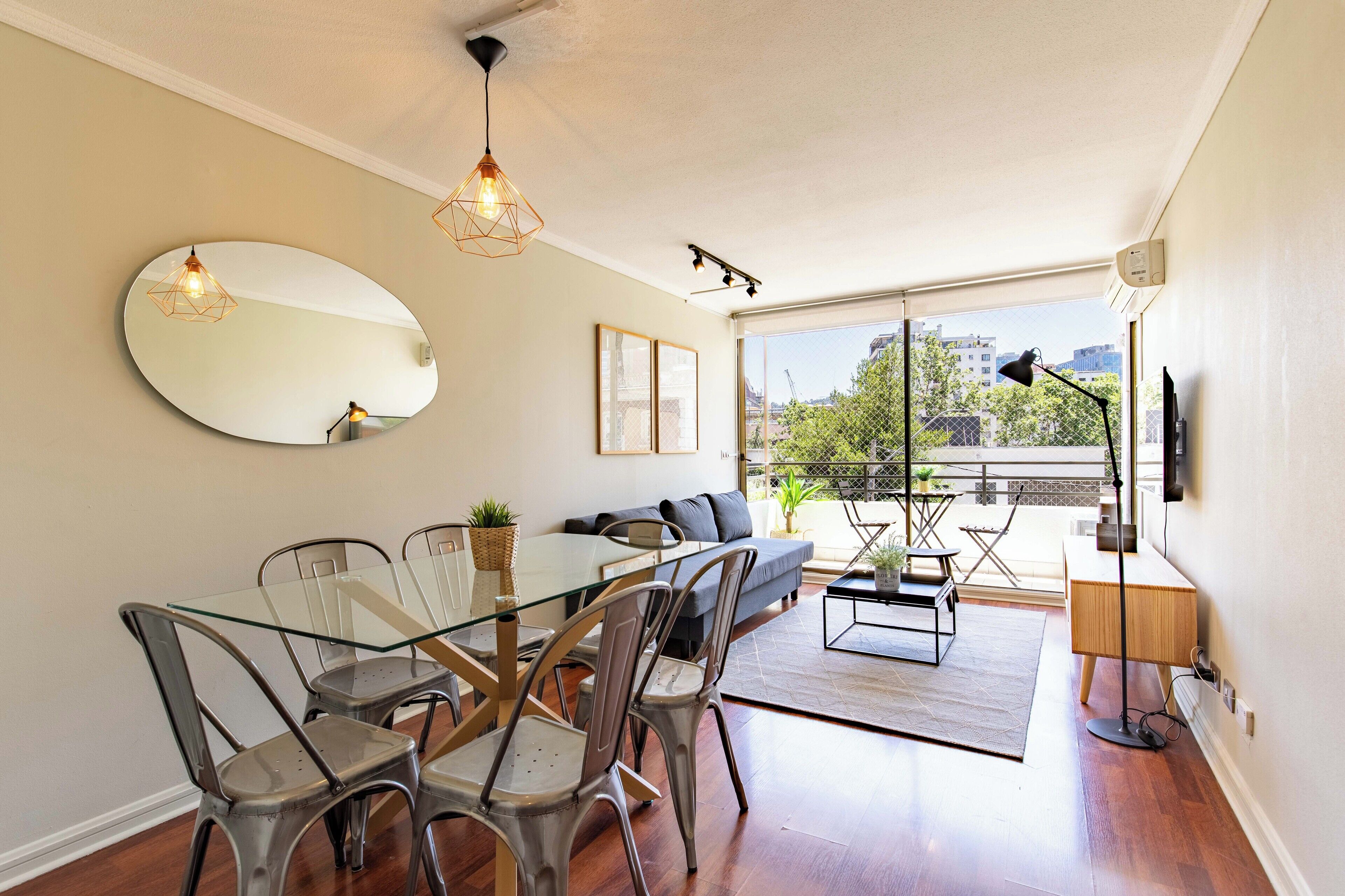 Un salon moderne avec une table à manger en verre, des chaises en métal, un canapé, un grand miroir et une vue sur la ville grâce à de grandes fenêtres.