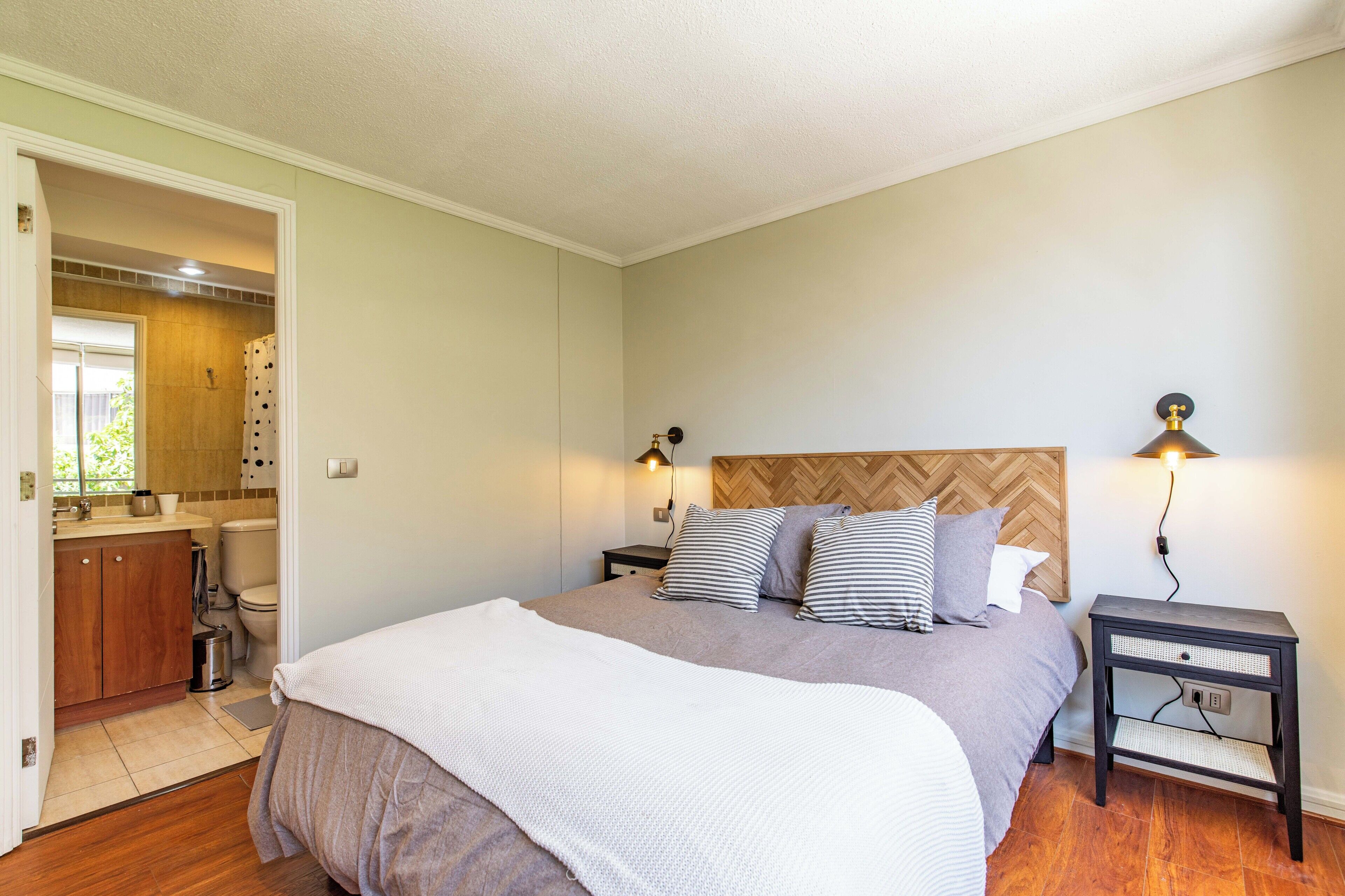 A bedroom with a bed, bedside tables, a nightstand, a wooden headboard, and a bathroom visible through an open door.