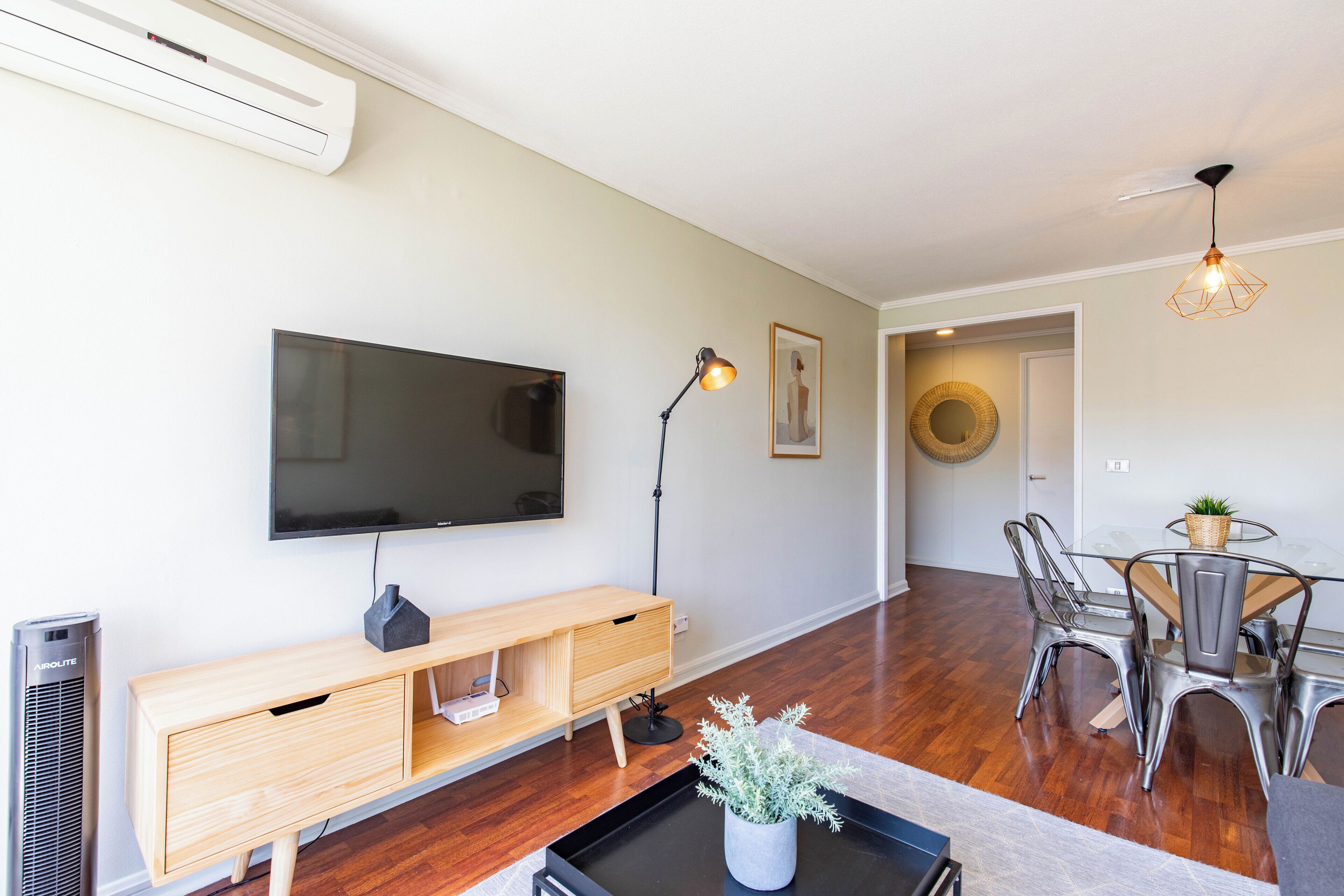 A modern living room with a flat-screen TV, wooden console, and a dining area with metal chairs and a glass table.