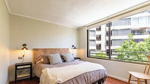 A bedroom with a bed, two bedside tables, a nightstand, a window with a view of buildings and greenery, and a chair. - 2-bedroom apartment in peaceful Providencia with AC and charm (Providencia)