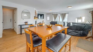 A spacious living area with a wooden dining table, black leather chairs, a black leather sofa, and a large window with curtains. - Walkable Adventure Base in Midtown Anchorage (Anchorage)