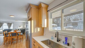 A kitchen with wooden cabinets, a stainless steel sink, and a window with a view of a snowy landscape. - Walkable Adventure Base in Midtown Anchorage (Anchorage)