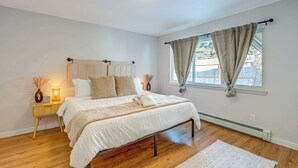 A bedroom with a bed, wooden flooring, a nightstand, a bedside lamp, and a window with curtains. - Walkable Adventure Base in Midtown Anchorage (Anchorage)