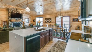 A cabin interior with a stone fireplace, wooden ceiling, a dining area, and a kitchen with a large island.