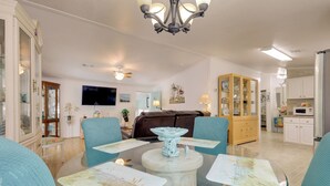 A spacious living room with a glass coffee table, blue upholstered chairs, a chandelier, and a television mounted on the wall.
