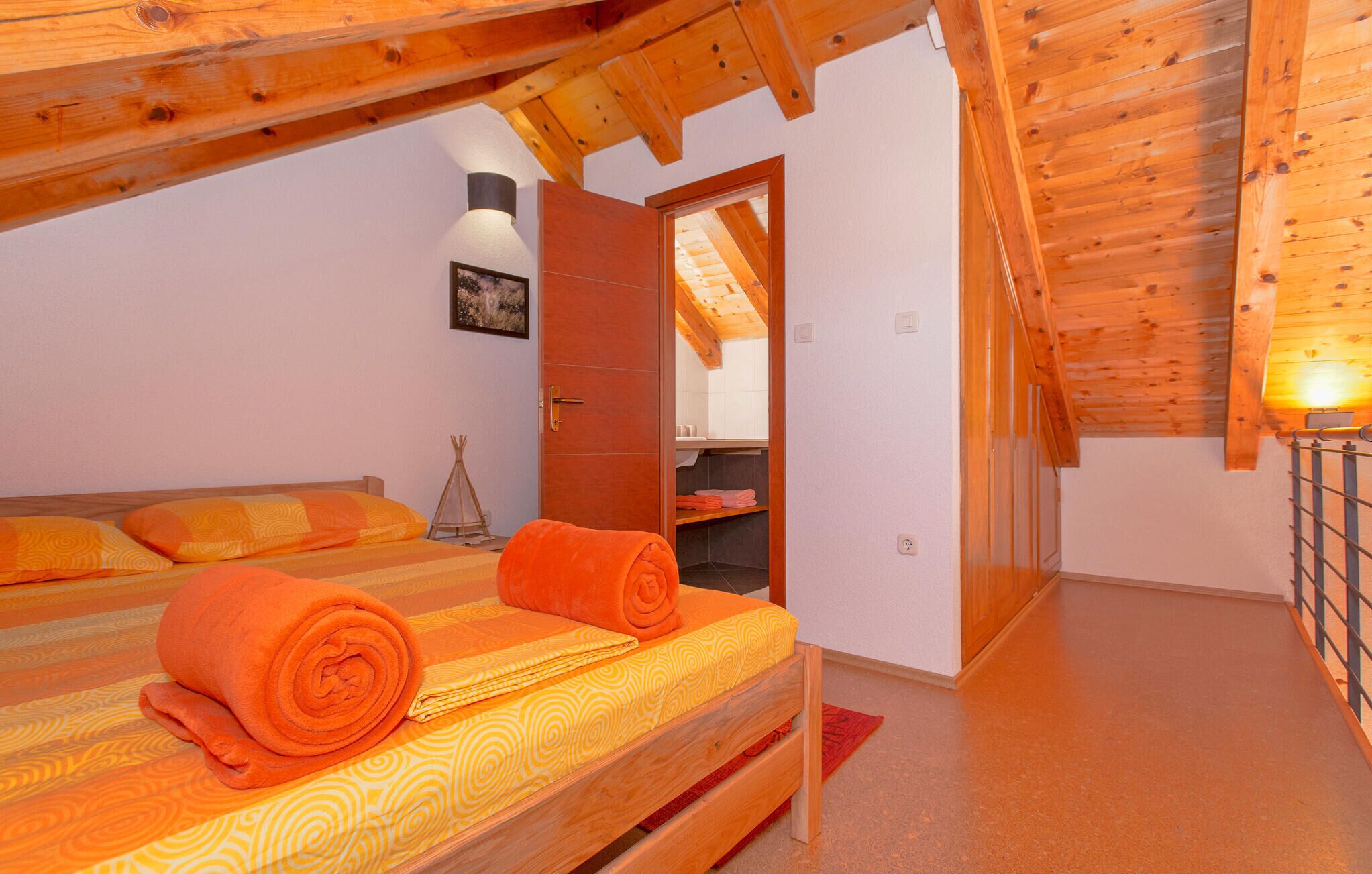 A cozy room with a wooden ceiling, a bed with orange bedding, a small window, and a door leading to a bathroom.