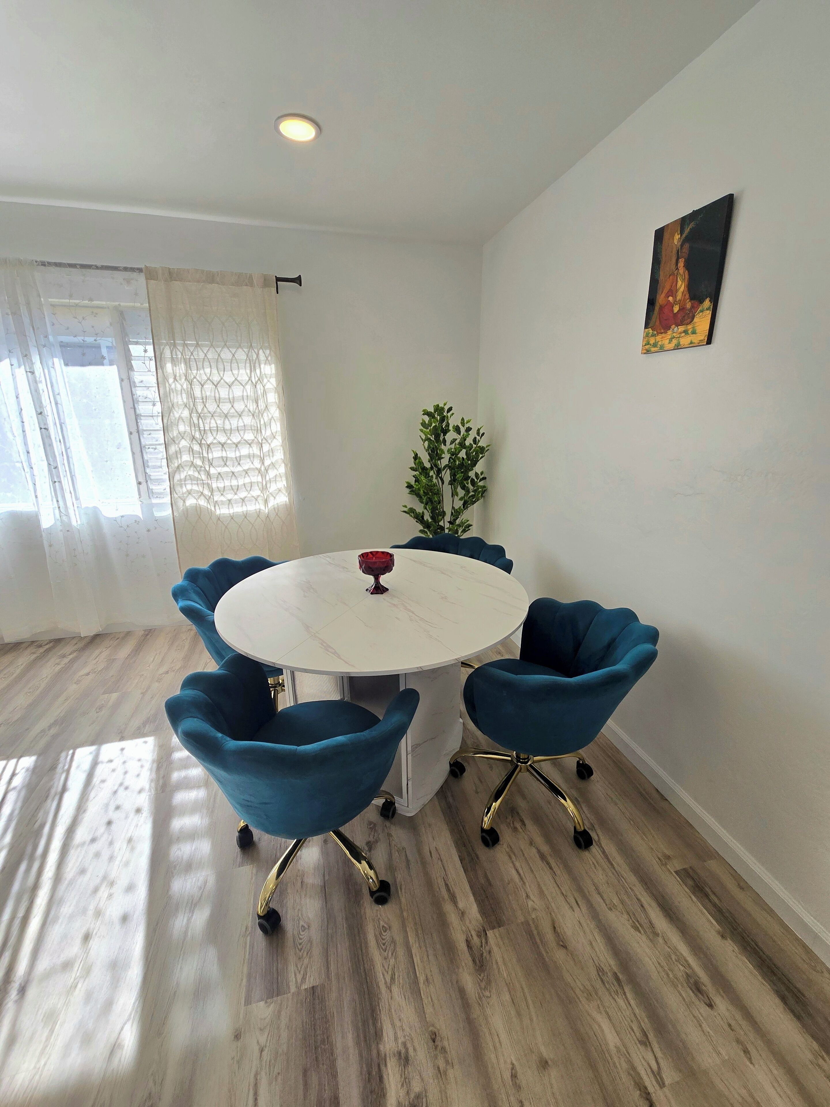 Una habitación con una mesa redonda blanca rodeada por cuatro sillas azules con patas doradas, una planta en maceta y un cuadro en la pared.