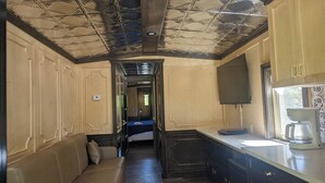 Un estrecho pasillo con una cama, un sofá, una zona de cocina con cafetera y un televisor.