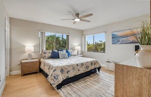 Une chambre à coucher comprenant un lit, des tables de chevet, un ventilateur de plafond, de grandes fenêtres et une vue sur l’extérieur.