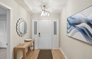 A hallway with a white door, a wooden console table with a bowl, a mirror with a circular frame, a chandelier, and a large abstract painting on the wall. - Walk to Beach & Dining | Pool + 2-Car Garage (Sarasota)