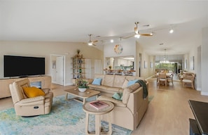 A spacious living room with a large flat-screen TV, a reclining chair, a sofa, and a dining area with a kitchen in the background. - Walk to Beach & Dining | Pool + 2-Car Garage (Sarasota)