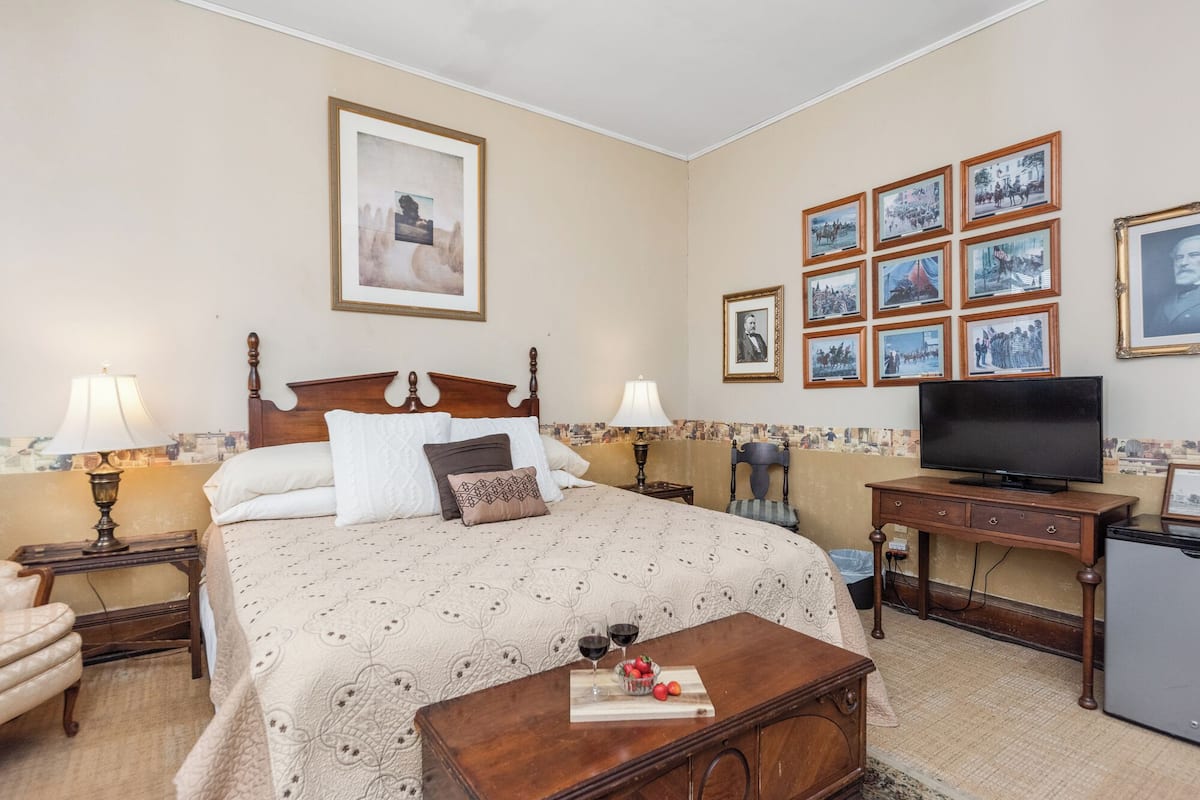 Habitación de hotel con una cama, dos mesitas de noche con lámparas, un televisor, un escritorio con una silla y una pared decorada con cuadros.