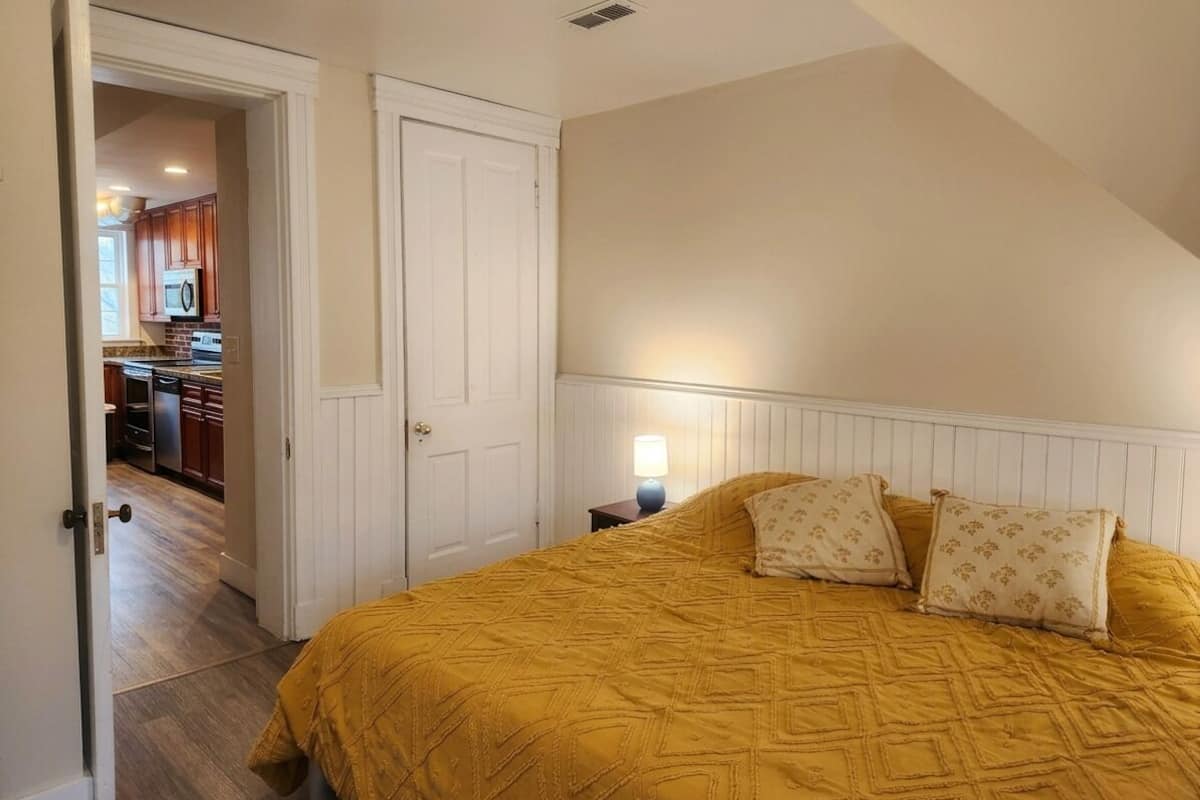 Un dormitorio con cama, mesita de noche, lámpara y puerta que da a una cocina.