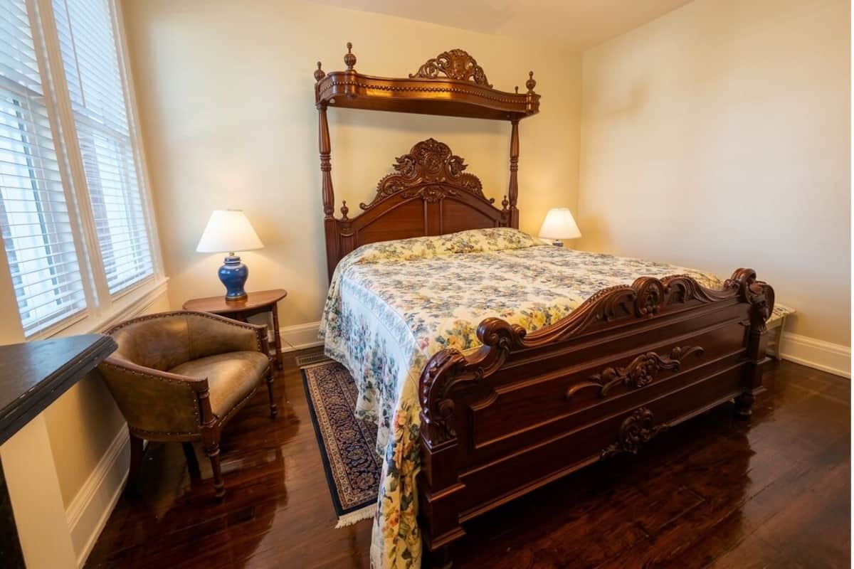 Un dormitorio con una cama con dosel, dos lámparas de noche, una silla y una ventana con persianas.