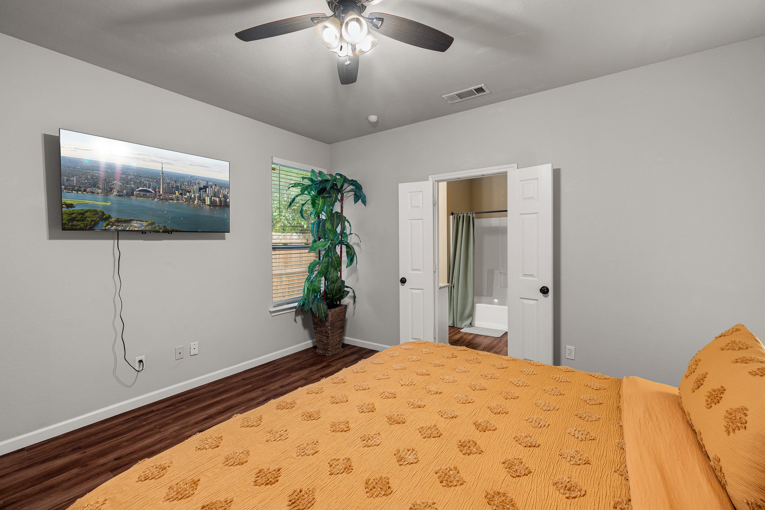 Un dormitorio con una cama, un ventilador de techo, un televisor, una planta en maceta y una puerta de baño.