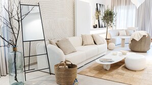 Uma sala de estar moderna com um sofá branco, uma mesa de centro e um espelho grande. Há uma planta em vaso e uma cesta trançada no chão.