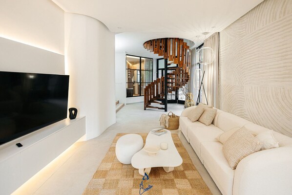 A modern living room with a flat-screen TV, a white sofa, a unique coffee table, and a staircase with wooden railings.
