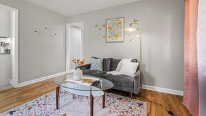 Ein Wohnzimmer mit Glastisch, grauem Sofa, einem floralen Teppich und einer Wandleuchte.
