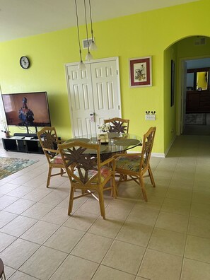 Un comedor con una mesa redonda de vidrio, cuatro sillas de madera con cojines coloridos, un televisor sobre un mueble, y una puerta blanca con un letrero rojo.