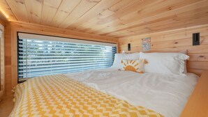 A wooden cabin bedroom with a bed, a window with blinds, and a wall-mounted light fixture.