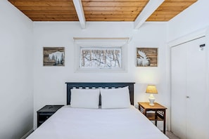 Un dormitorio con techo de madera, una cama con ropa de cama blanca, dos mesitas de noche y obras de arte en la pared que representan escenas naturales.