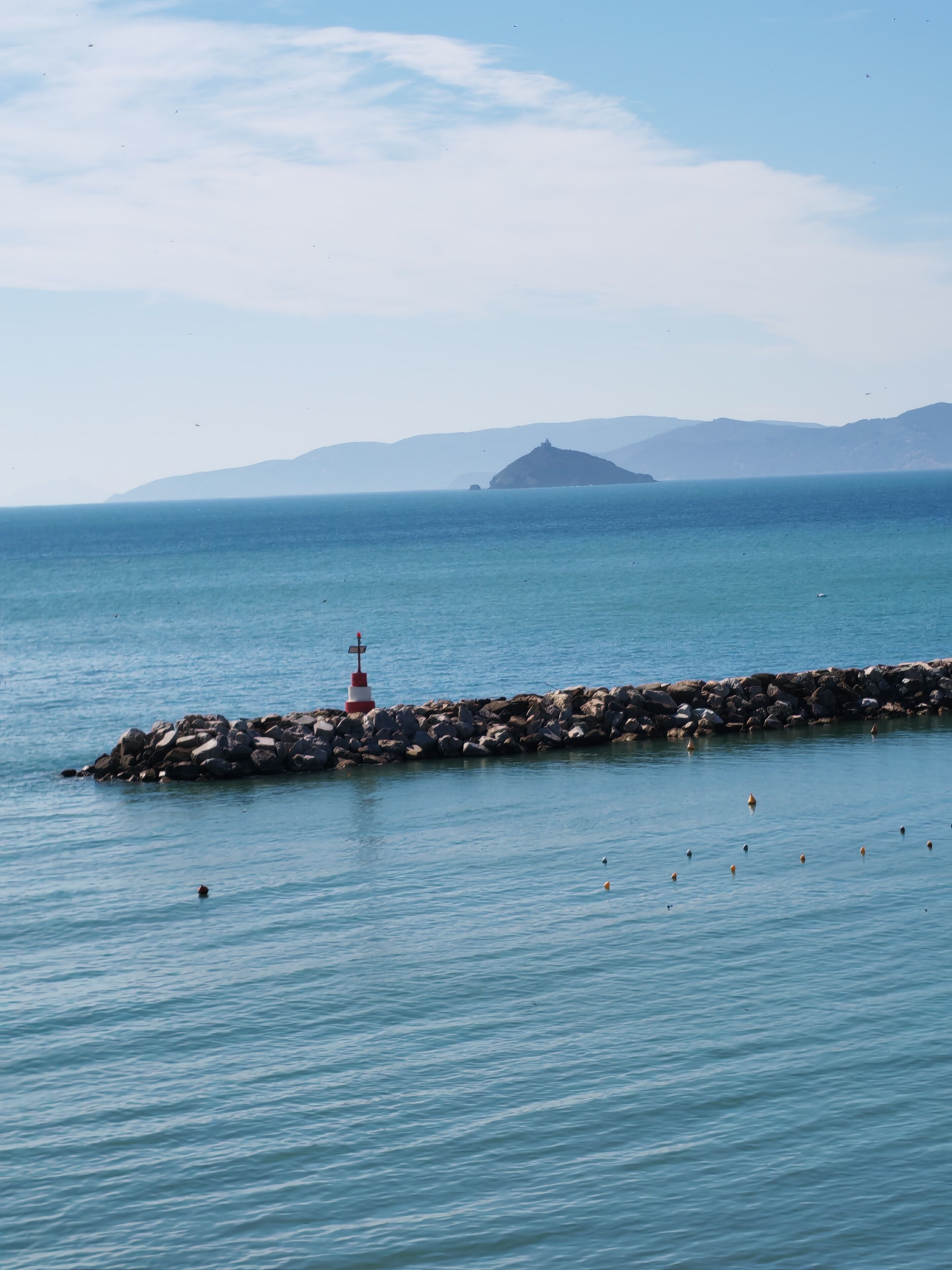 A lighthouse on a rocky breakwater with calm sea and distant mountains.