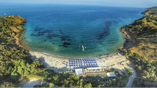 Une vue aérienne d’une plage aux eaux cristallines, bordée de falaises rocheuses et d’une rangée de parasols.
