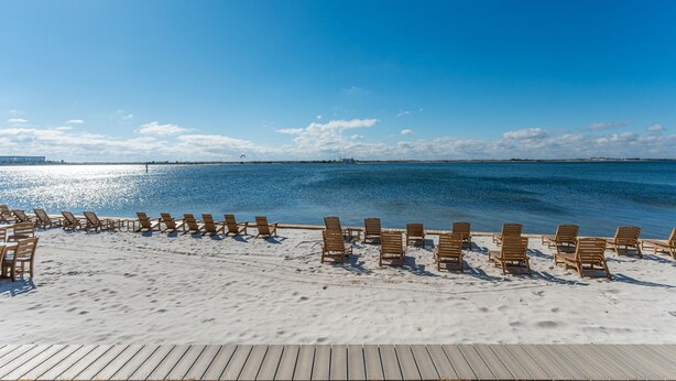 A beach with wooden lounge chairs arranged in rows on the sand, a wooden deck in the foreground, and a calm sea under a clear sky.
