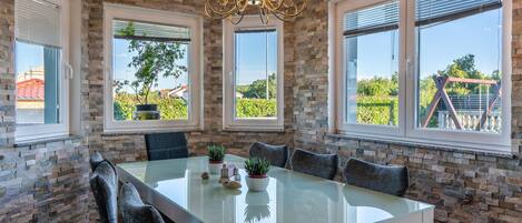 A dining area with a glass table, stone walls, and large windows with blinds.
