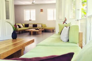 Un salon avec un canapé vert, une table basse en bois et un banc. Il y a des fenêtres avec des volets et un ventilateur de plafond.
