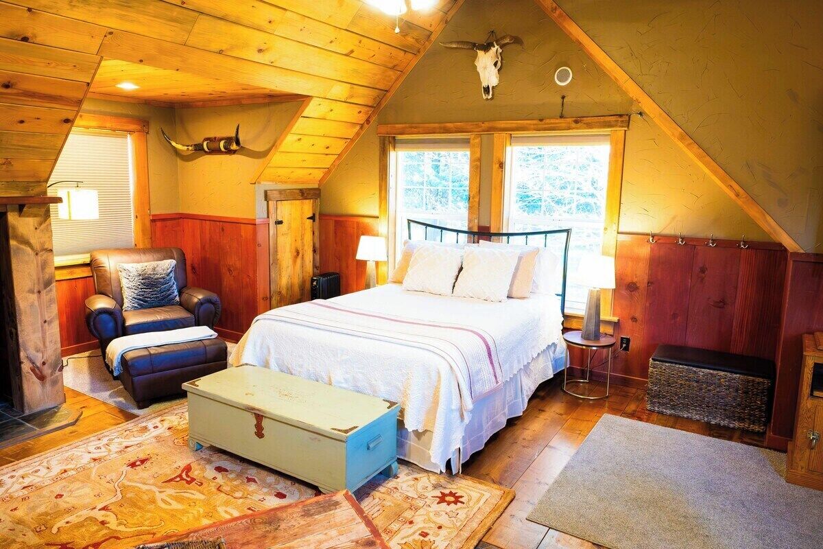 A cozy bedroom with a wooden ceiling, a bed with white bedding, a leather chair, and a rustic decor including a cow skull.