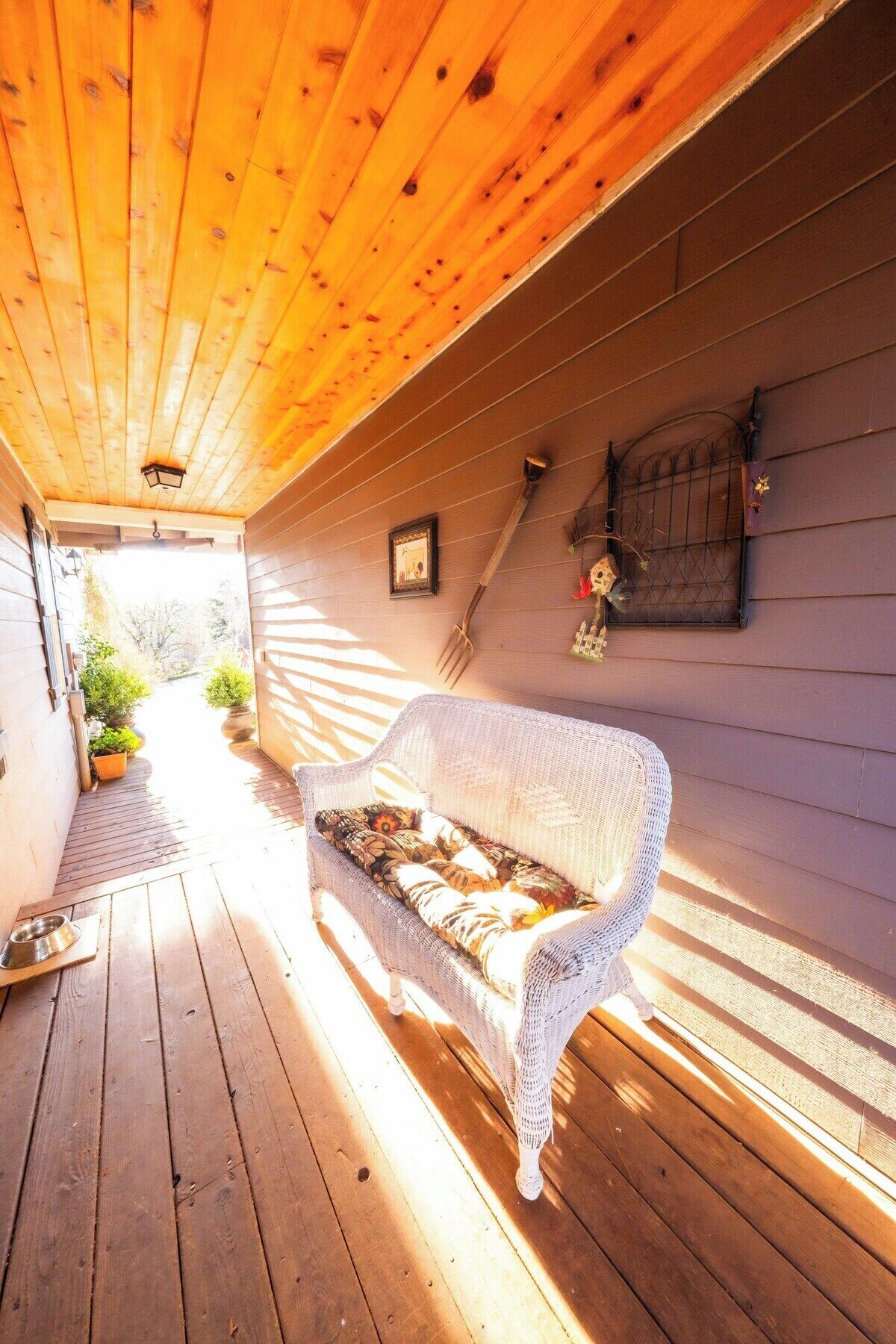 A covered porch with a wicker sofa, wooden floor, and a small garden area with potted plants.