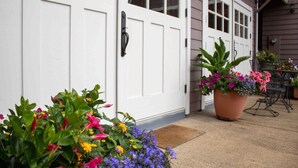 Une maison avec une porte d’entrée blanche, un porche brun et des plantes en pot ornées de fleurs.