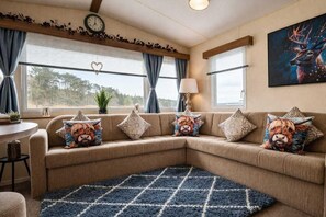 A living room with a sectional sofa, a blue rug, a clock, and a painting of a deer. - Cabin- Sleeps 8- Hot Tub, Free Parking, Wifi, Pets (Newton Stewart)