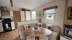 Una cocina compacta con estufa, horno y microondas. Hay una mesa redonda de madera con un vaso de agua y un plato de comida. Un sofá color beige con cojines de temática canina está apoyado en la pared.