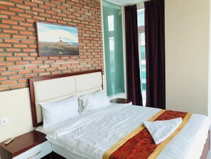 A hotel room with a brick wall, a bed with white bedding, a red and gold bedspread, a nightstand, and a window with curtains. - Sai&su hotel (Phnom Penh)