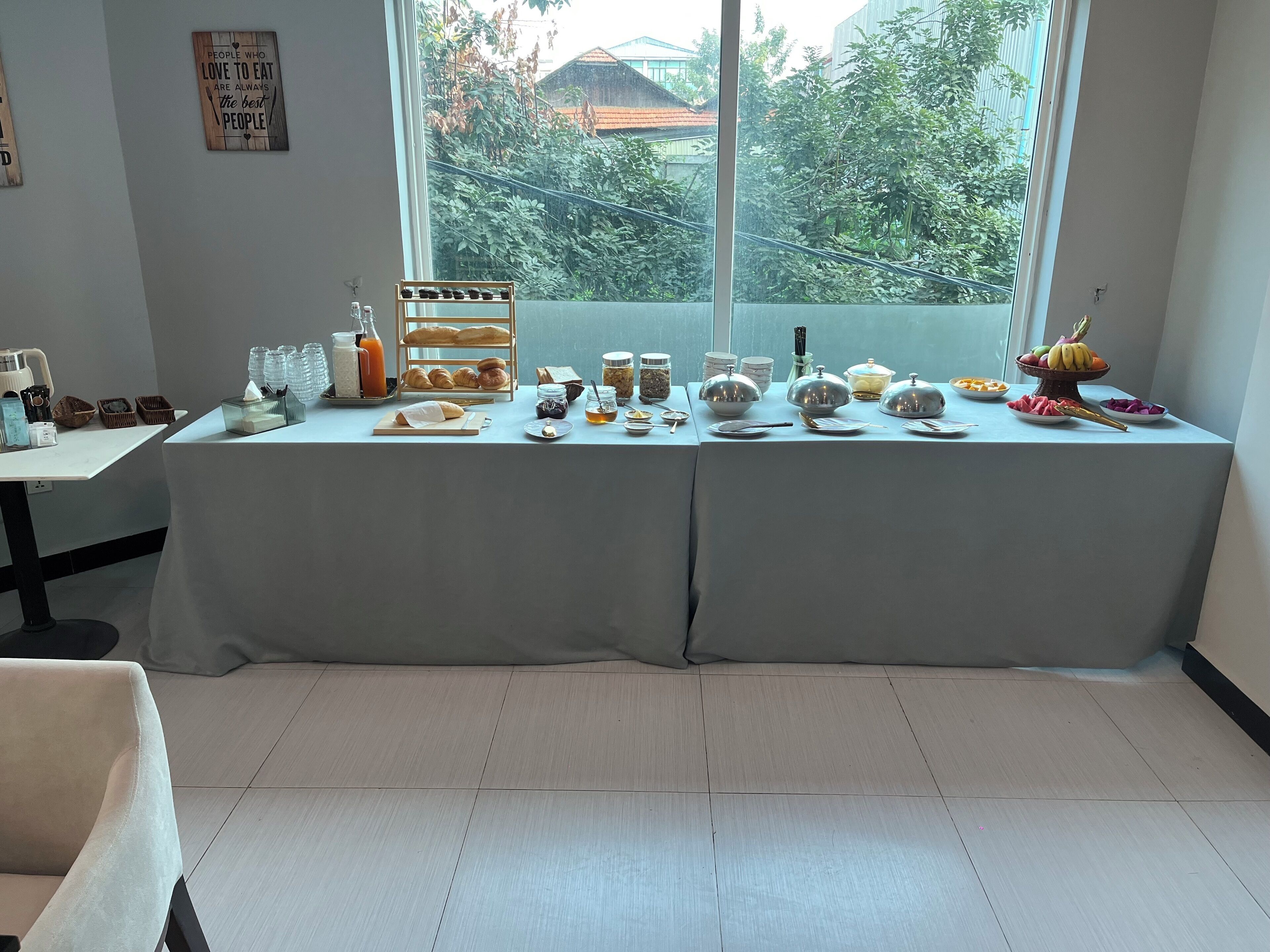 Ein reichhaltiges Frühstücksbuffet mit verschiedenen Speisen und Beilagen auf einem Tisch, ein Fenster mit Blick auf Grünflächen und eine Wand mit einem motivierenden Zitat.