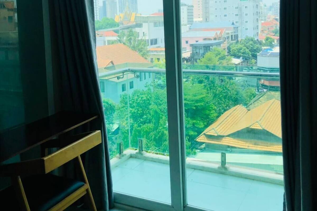 A room with a large window, a wooden chair, and a view of buildings and greenery outside.