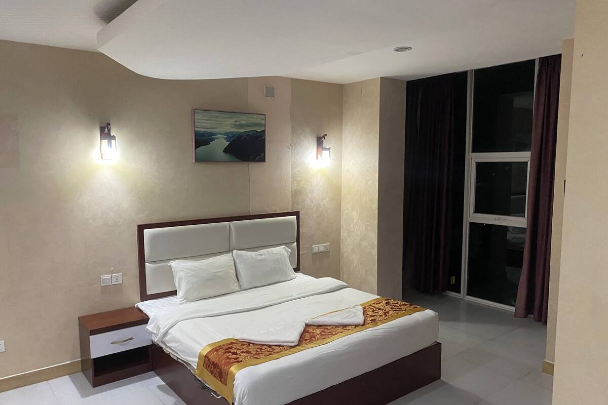 A modern hotel room with a large bed, nightstands, a painting on the wall, and a view through a window.