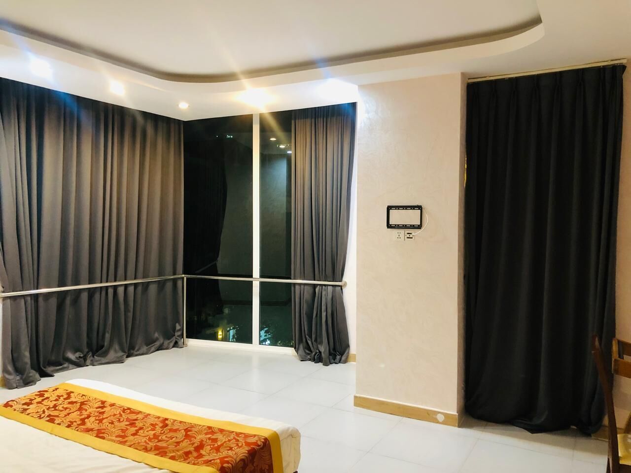 Una habitación de hotel con un ventanal grande, cortinas oscuras, una cama con colcha estampada y una mesita pequeña.