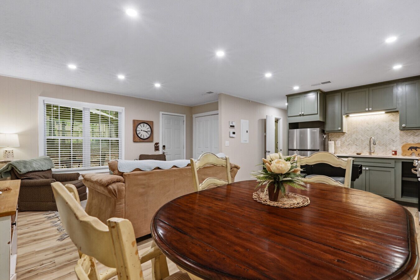 A dining area with a wooden table, beige upholstered chairs, a clock on the wall, and a kitchen with grey cabinets and stainless steel appliances.