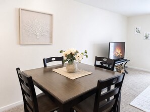 Un coin repas comprenant une table, des chaises, un vase avec des fleurs, un tableau encadré au mur et un téléviseur diffusant une image de cheminée.