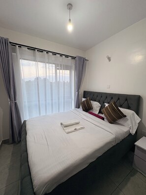 A hotel room with a bed, curtains, a bedside table, and a wall-mounted light fixture. - Cozy &stylish clean one bedroom Apartment (Ongata Rongai)