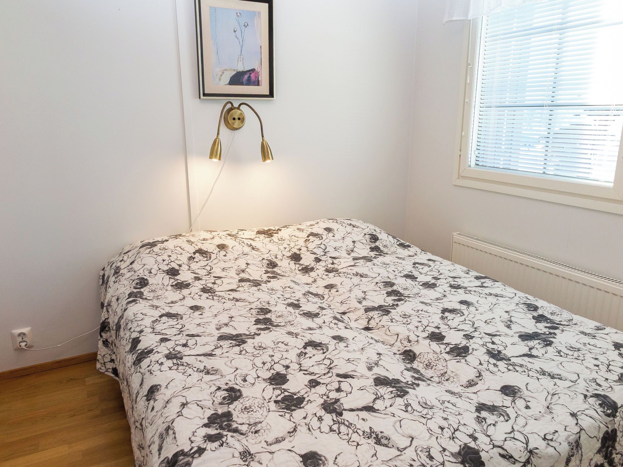 Un dormitorio con colcha de estampado floral, lámpara de pared y ventana con persianas.